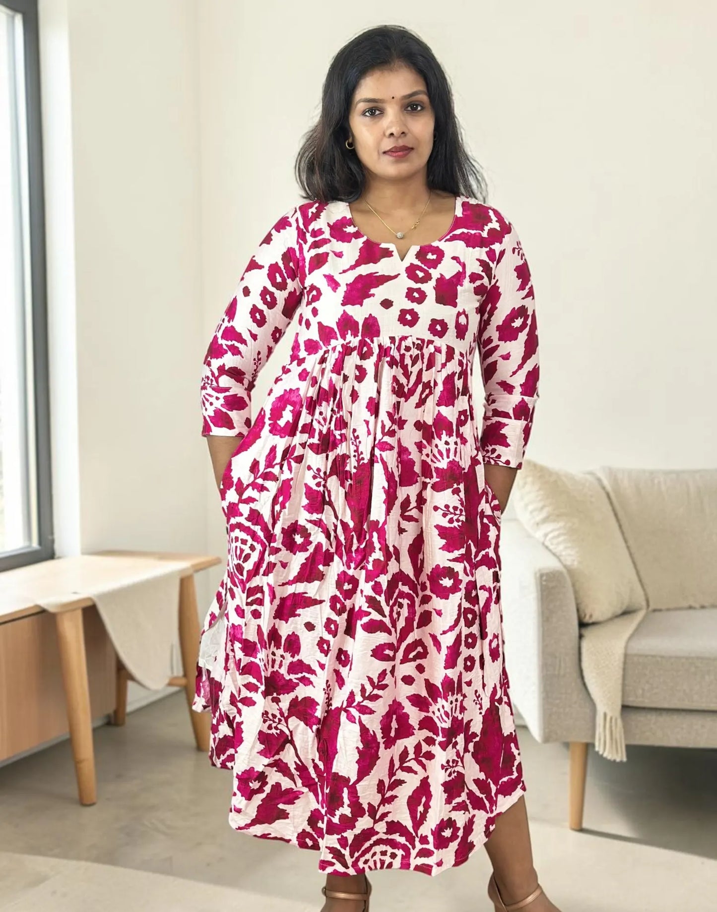 A woman wearing a long-sleeved midi dress with a bold magenta and white floral pattern, standing in a brightly lit, modern living room.
