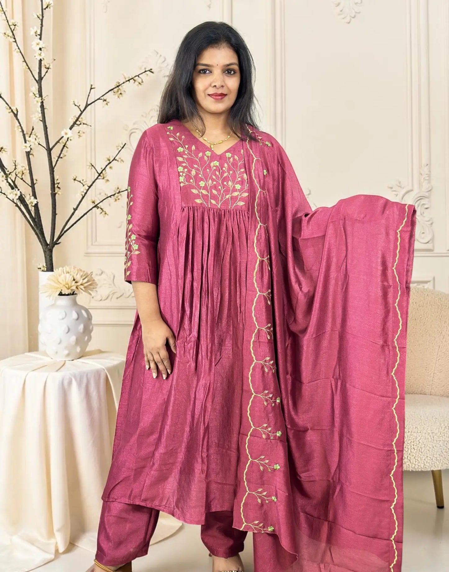 A woman models a traditional Indian salwar kameez suit in a dusty rose pink color with gold embroidery, paired with a matching dupatta, set against an elegant cream-colored interior background.