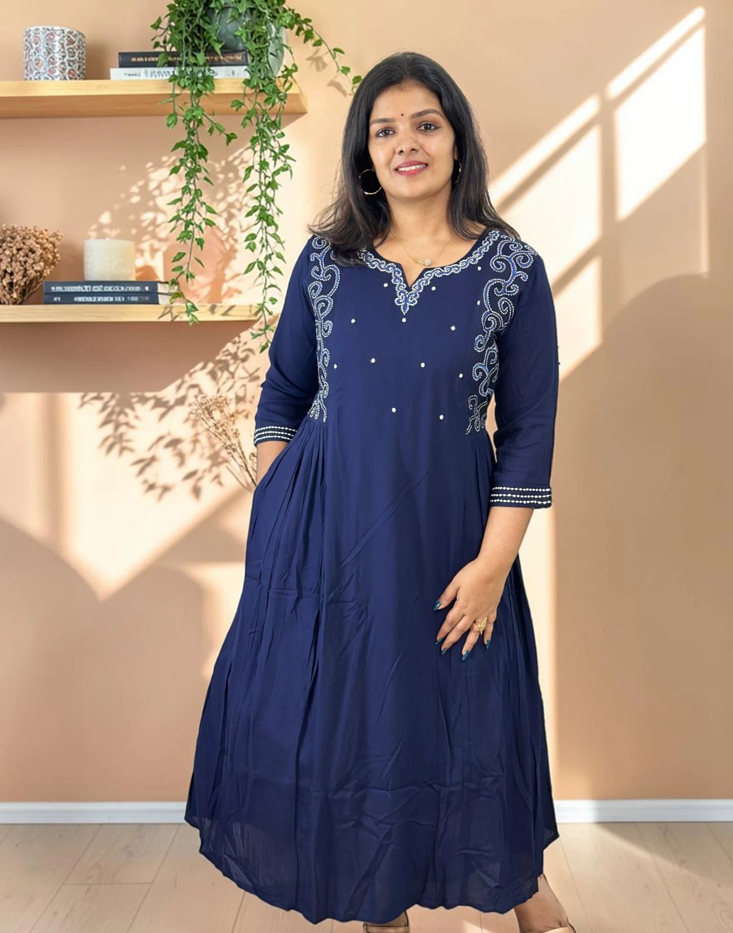 A woman modeling a navy blue A-line kurta with silver embroidery on the neckline and sleeve cuffs, standing in a warm-toned room with sunlight casting shadows on the wall.