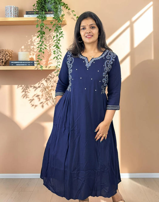 A woman modeling a navy blue A-line kurta with silver embroidery on the neckline and sleeve cuffs, standing in a warm-toned room with sunlight casting shadows on the wall.