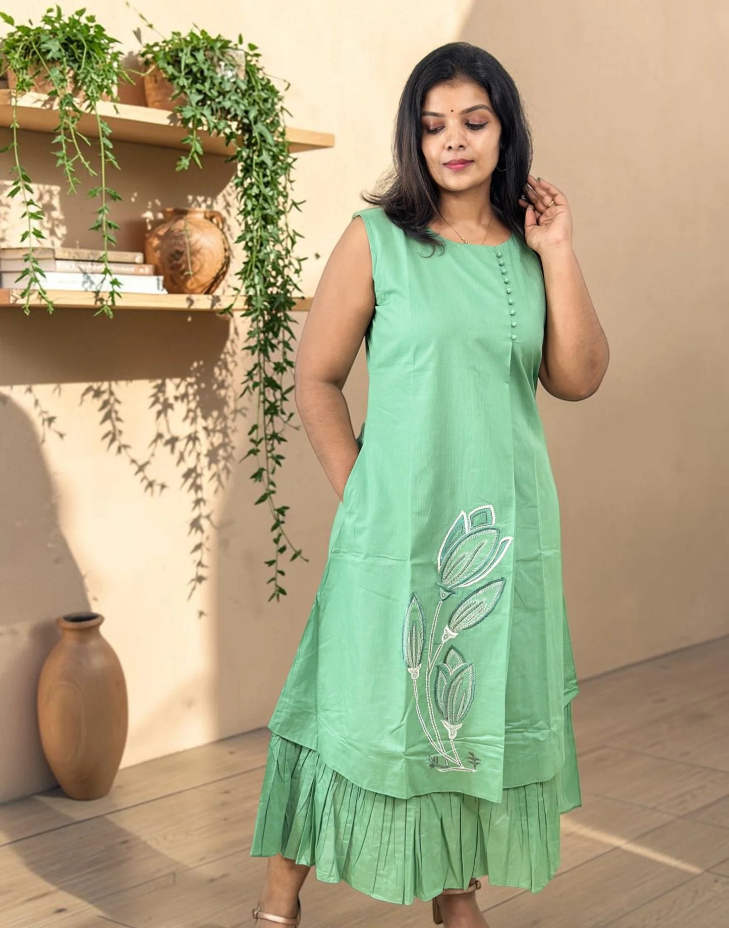 A woman wearing a sleeveless sage green kurta with white floral embroidery and a tiered ruffled hem, standing in a warm-toned indoor setting with shelves and plants in the background.