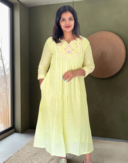 A woman wearing a light yellow-green A-line kurta with pink floral embroidery on the chest, standing in a room with a dark olive green wall and a woven wall hanging.