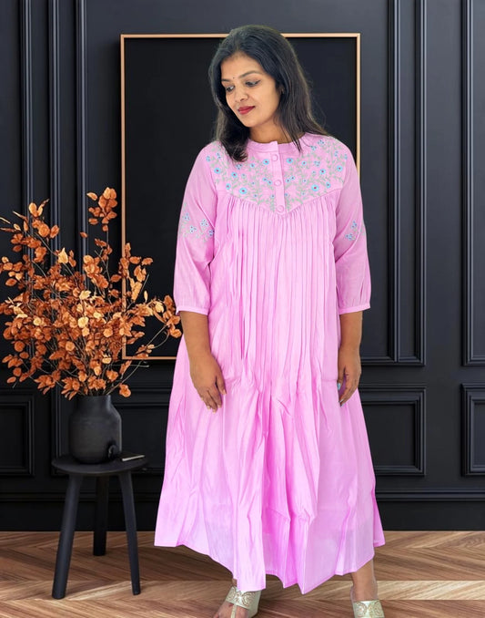A woman wearing a light pink tiered cotton kurta with floral embroidery on the yoke and sleeves, standing in front of a dark paneled wall.