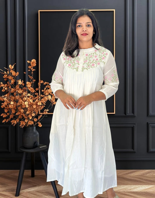 A woman wearing a white midi dress with pink and green floral embroidery on the chest and sleeves, standing in front of a black paneled wall with a gold frame.