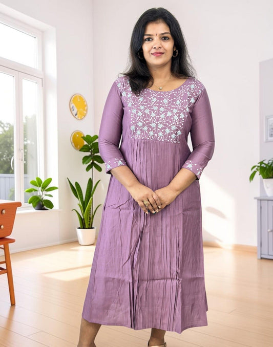 A woman wearing a lavender purple three-quarter sleeve kurta with white floral embroidery on the chest, standing in a bright, modern room with light wood floors and potted plants.