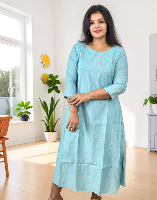 A woman wearing a light blue cotton kurta with pink floral embroidery on the chest, standing in a bright, modern room with plants and a window.