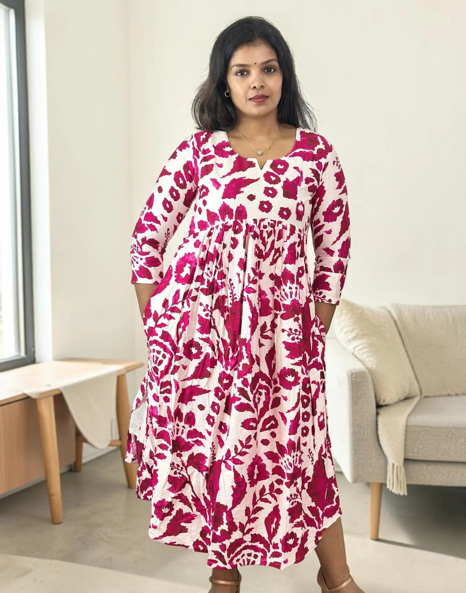 A woman wearing a long-sleeved midi dress with a bold magenta and white floral pattern, standing in a brightly lit, modern living room.
