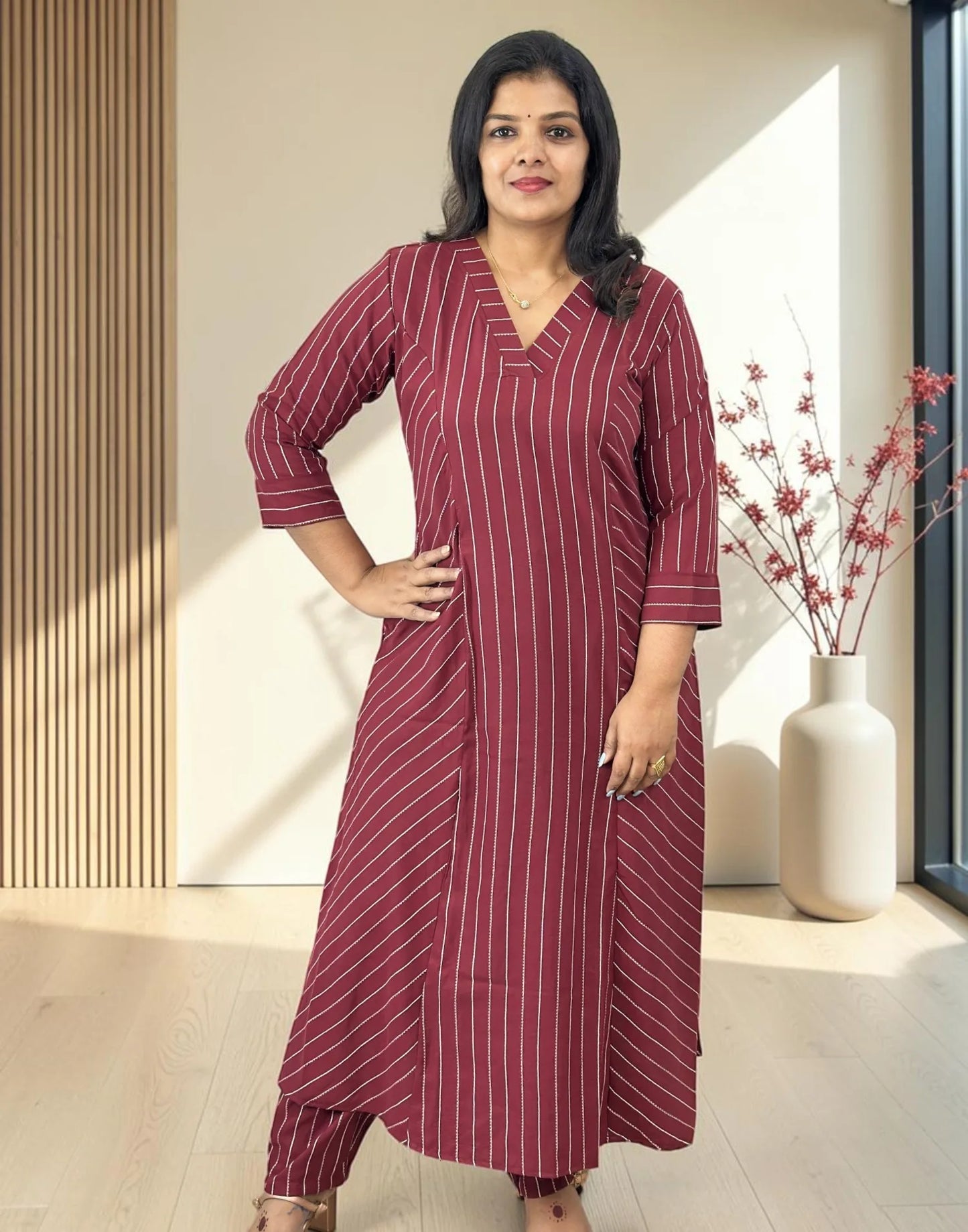 A woman modeling a long-sleeved, V-neck kurta in maroon with thin white vertical stripes, standing in a modern indoor setting with a beige wall, wooden slats, and a vase of branches.