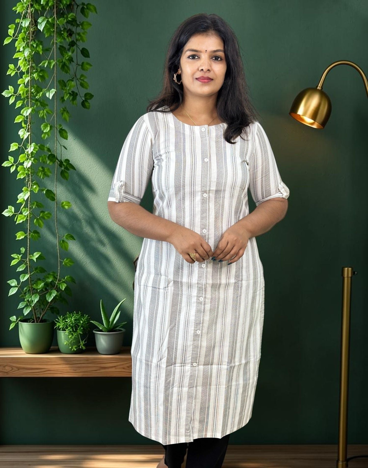 A woman wearing a white and gray vertically striped short-sleeve tunic dress with a button-down front, standing in front of a dark green wall with a gold floor lamp and potted plants.
