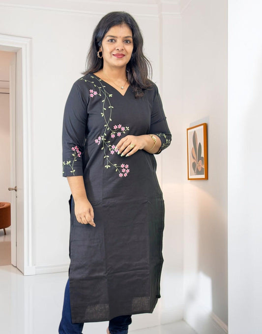 A woman modeling a black cotton kurta with pink floral embroidery on the chest and sleeve, paired with blue jeans in a bright indoor hallway.