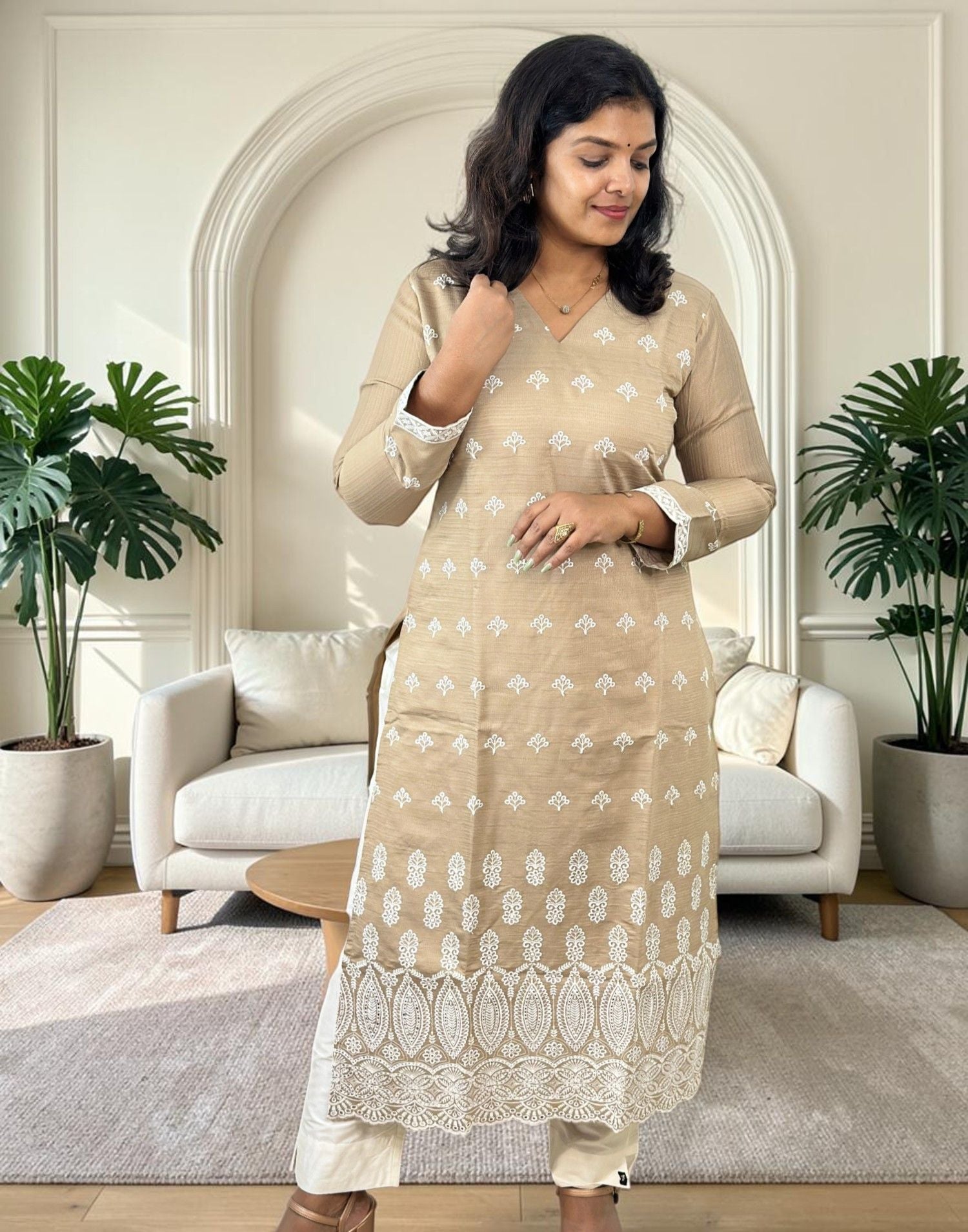 A woman modeling a beige kurta with white printed motifs and a white lace hem, paired with white pants in a modern living room setting.