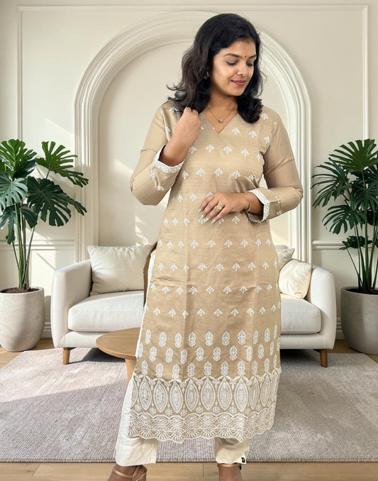 A woman modeling a beige kurta with white printed motifs and a white lace hem, paired with white pants in a modern living room setting.