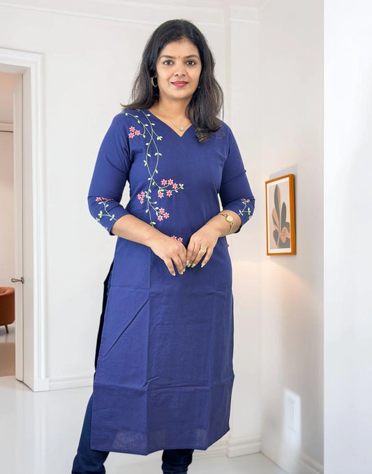 A woman wearing a royal blue cotton kurta with pink and green floral embroidery on the chest and sleeves, standing in a bright white room.