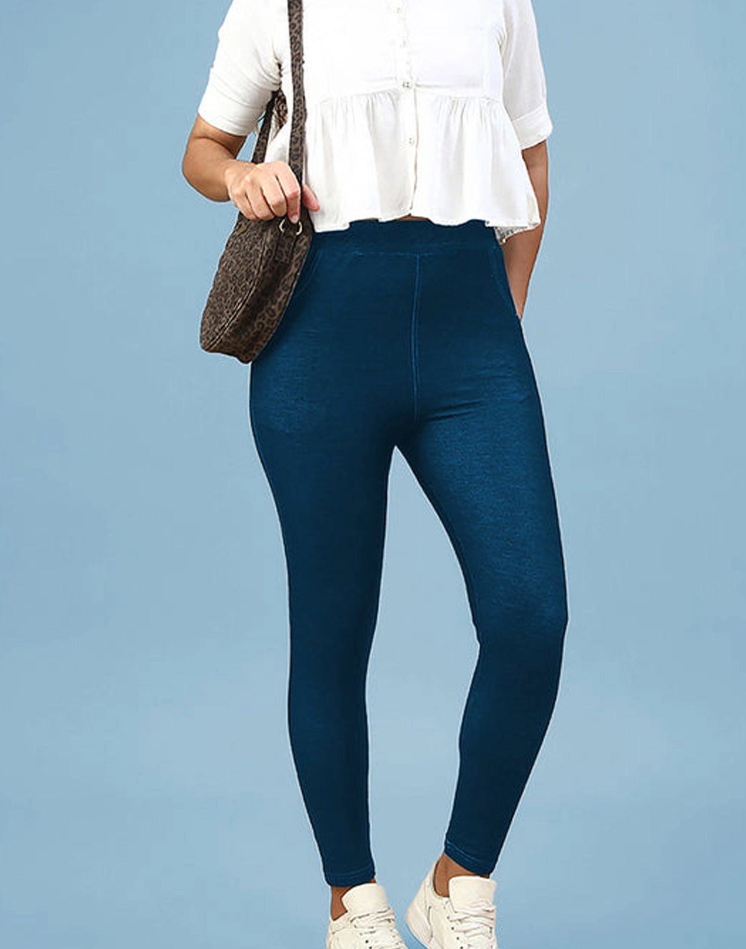 A model wearing dark blue high-waisted leggings, a white short-sleeve peplum blouse, and white sneakers, carrying a brown textured shoulder bag against a solid light blue background.