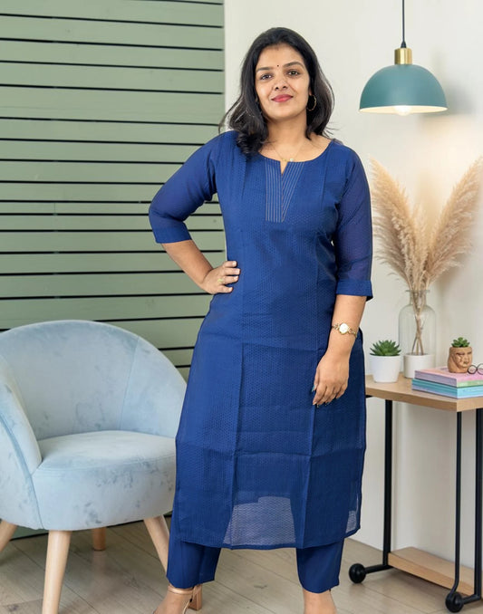 A woman modeling a blue three-quarter sleeve kurta with matching pants in a styled indoor setting with a light blue chair, a green slatted wall, and a side table with decorative items.