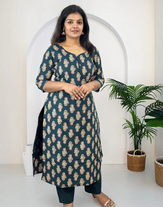 A woman modeling a dark teal kurta with beige and gold floral motifs, paired with dark pants, standing in front of a white arched wall with a potted plant to the side.