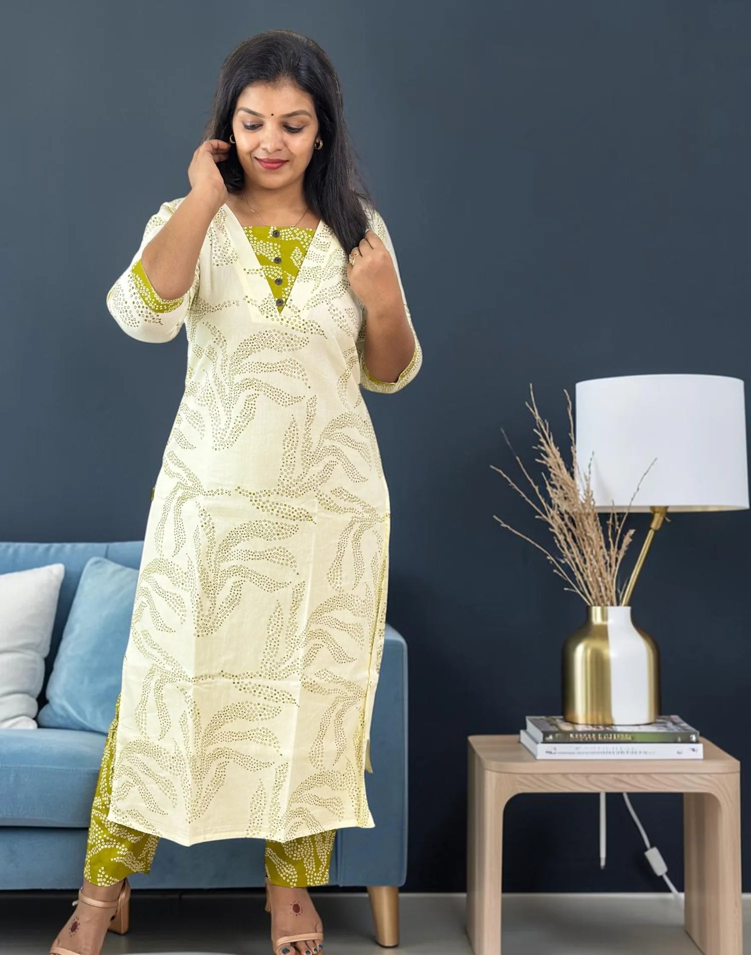 A woman modeling a cream-colored kurta with a green and beige leaf pattern, paired with matching patterned pants, standing in a living room setting next to a blue sofa and a side table with a lamp.