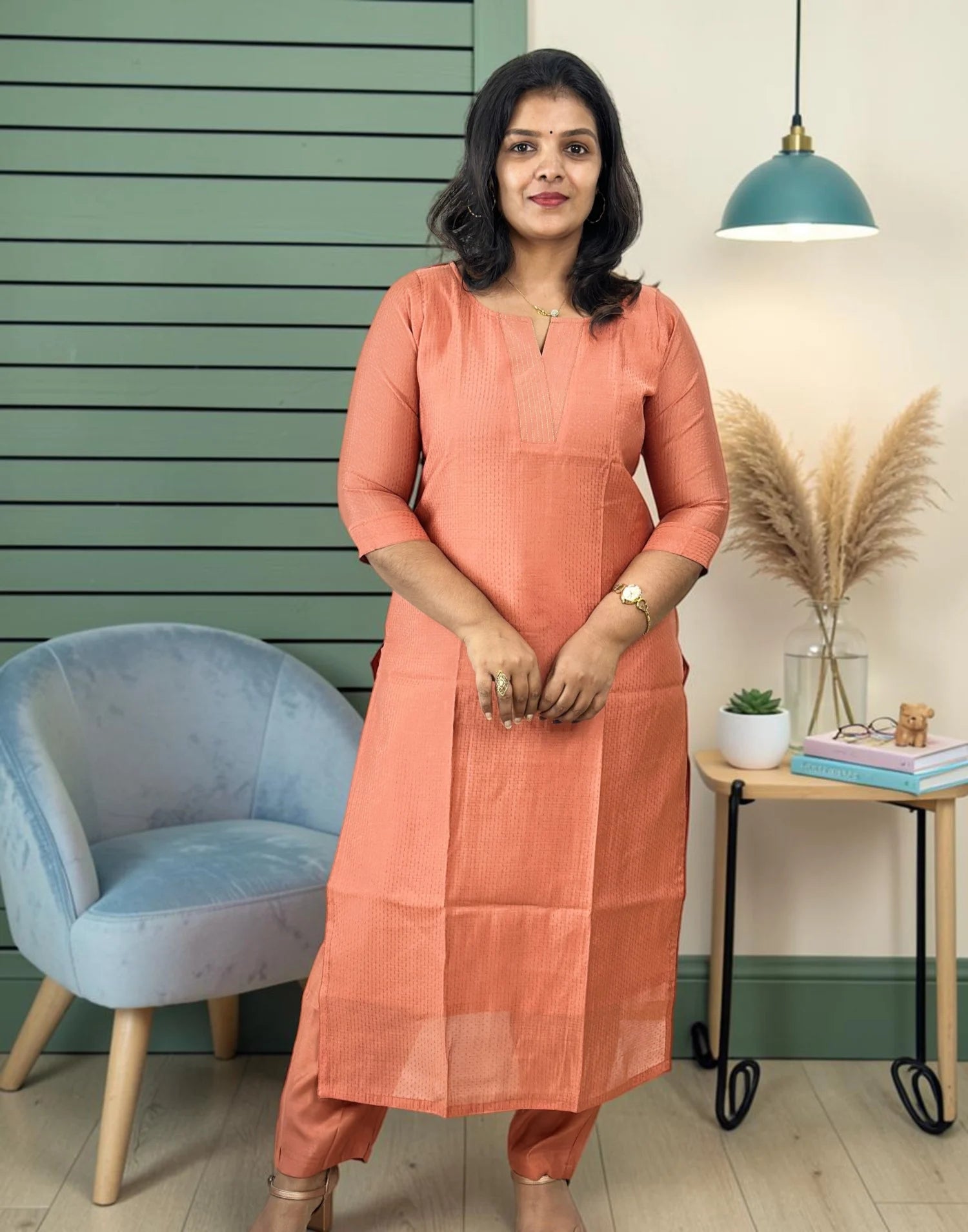 A woman wearing a peach-colored cotton kurta and matching trousers stands in a styled room with a green slatted wall, a blue armchair, and a side table with books and pampas grass.