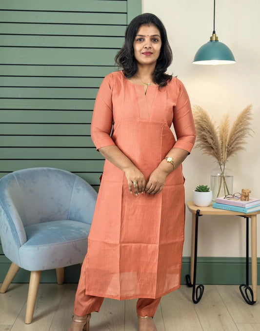 A woman wearing a peach-colored cotton kurta and matching trousers stands in a styled room with a green slatted wall, a blue armchair, and a side table with books and pampas grass.
