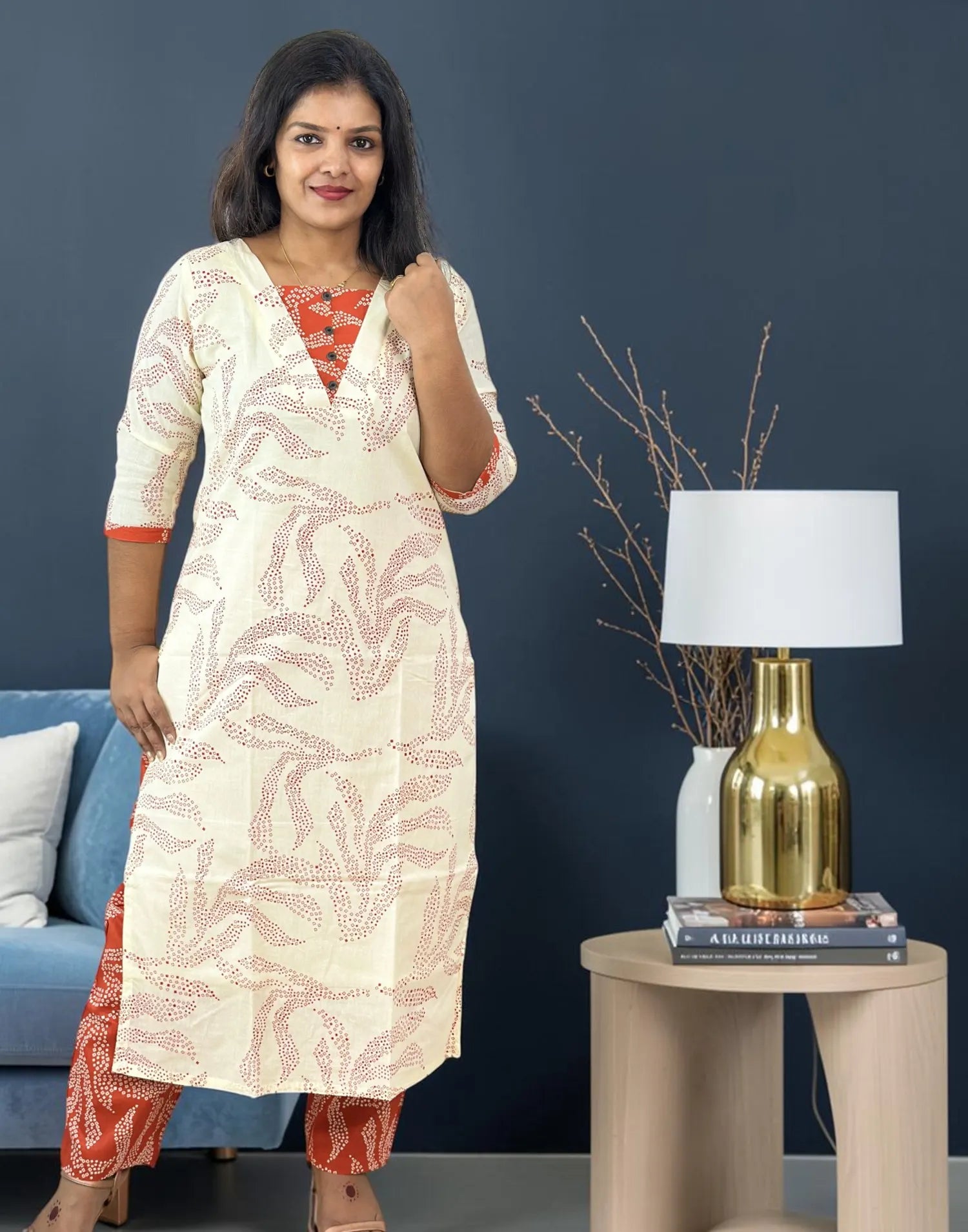 A woman models a cream-colored kurta with a dusty pink leaf pattern, paired with orange patterned pants, standing in a styled room next to a side table with a lamp.