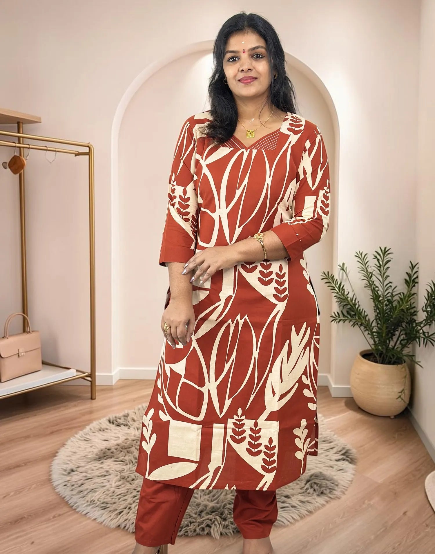 A woman modeling a rust-red and cream printed kurta with matching pants in a styled indoor boutique setting.