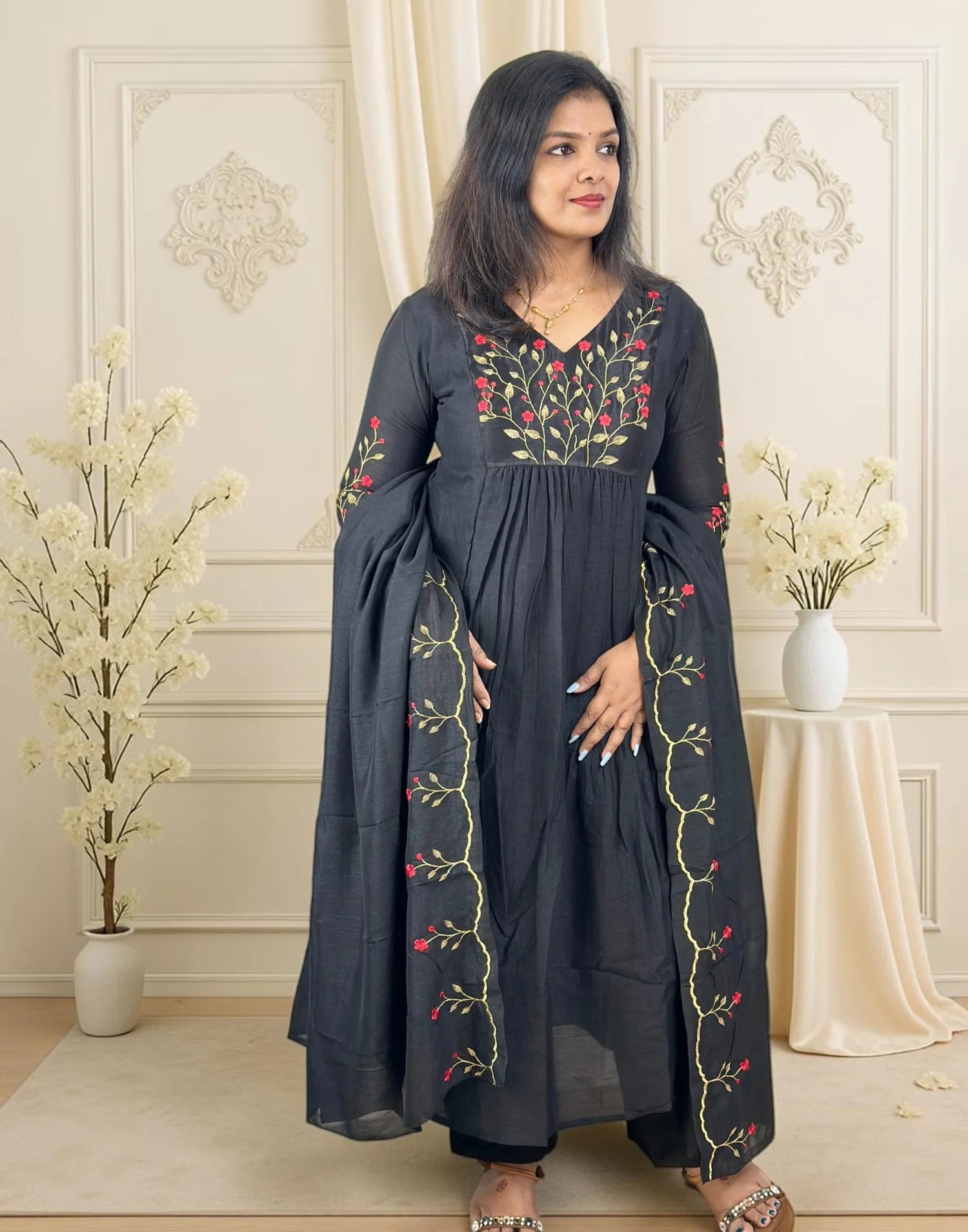 A woman modeling a dark charcoal gray long kurta with colorful floral embroidery in red, pink, and gold, set against an elegant cream-colored paneled wall background with decorative dried flowers.