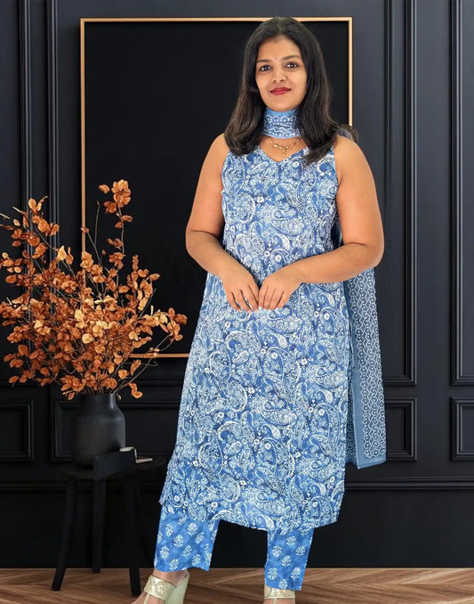 A woman modeling a blue and white printed salwar kameez suit with a matching dupatta, standing in front of a dark paneled wall with a decorative plant on the left.