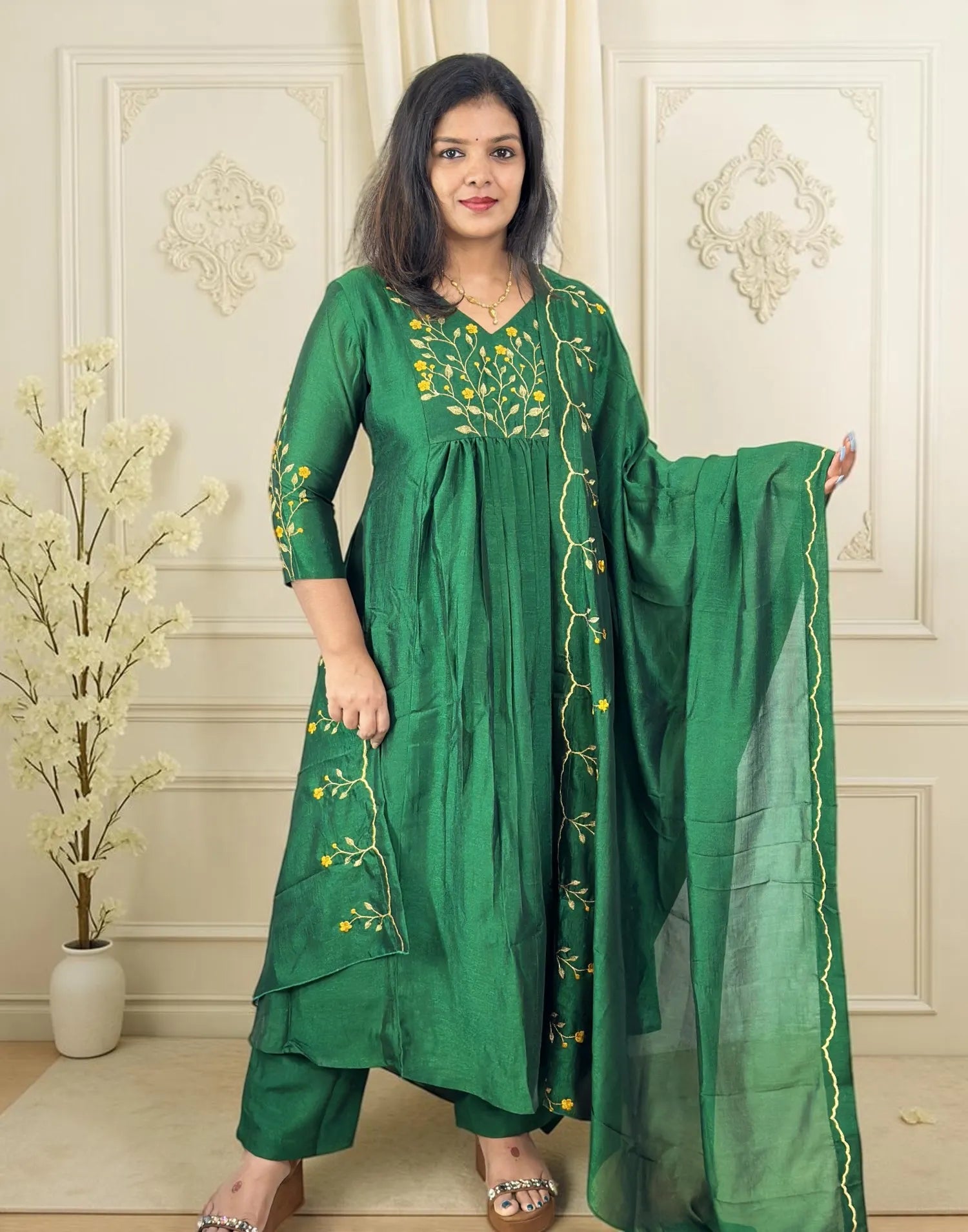 A woman models a green traditional Indian salwar kameez suit with yellow floral embroidery and a matching sheer green dupatta, set against an elegant cream-colored paneled wall.