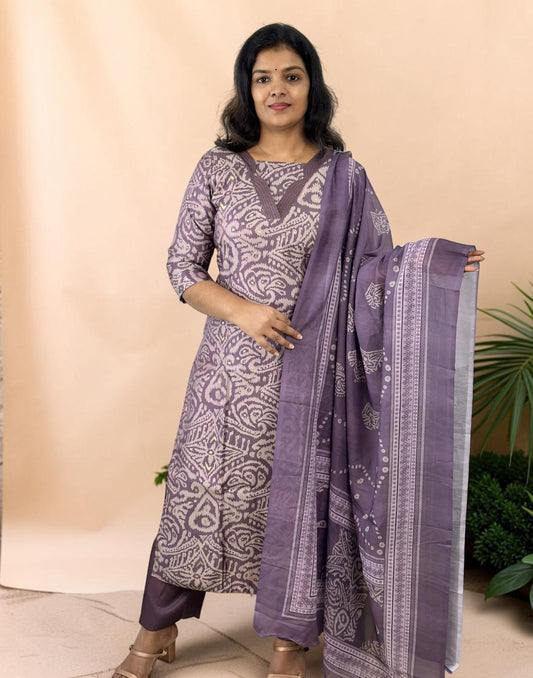 A woman modeling a three-piece traditional Indian salwar kameez set in lavender purple with white block prints, featuring a kurta, matching trousers, and a printed dupatta, set against a solid beige backdrop.