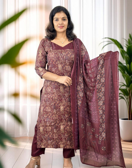 A woman modeling a traditional Indian salwar kameez suit in a dusty rose and gold floral print, paired with matching trousers and a sheer dupatta, set against a softly lit indoor background with potted plants.