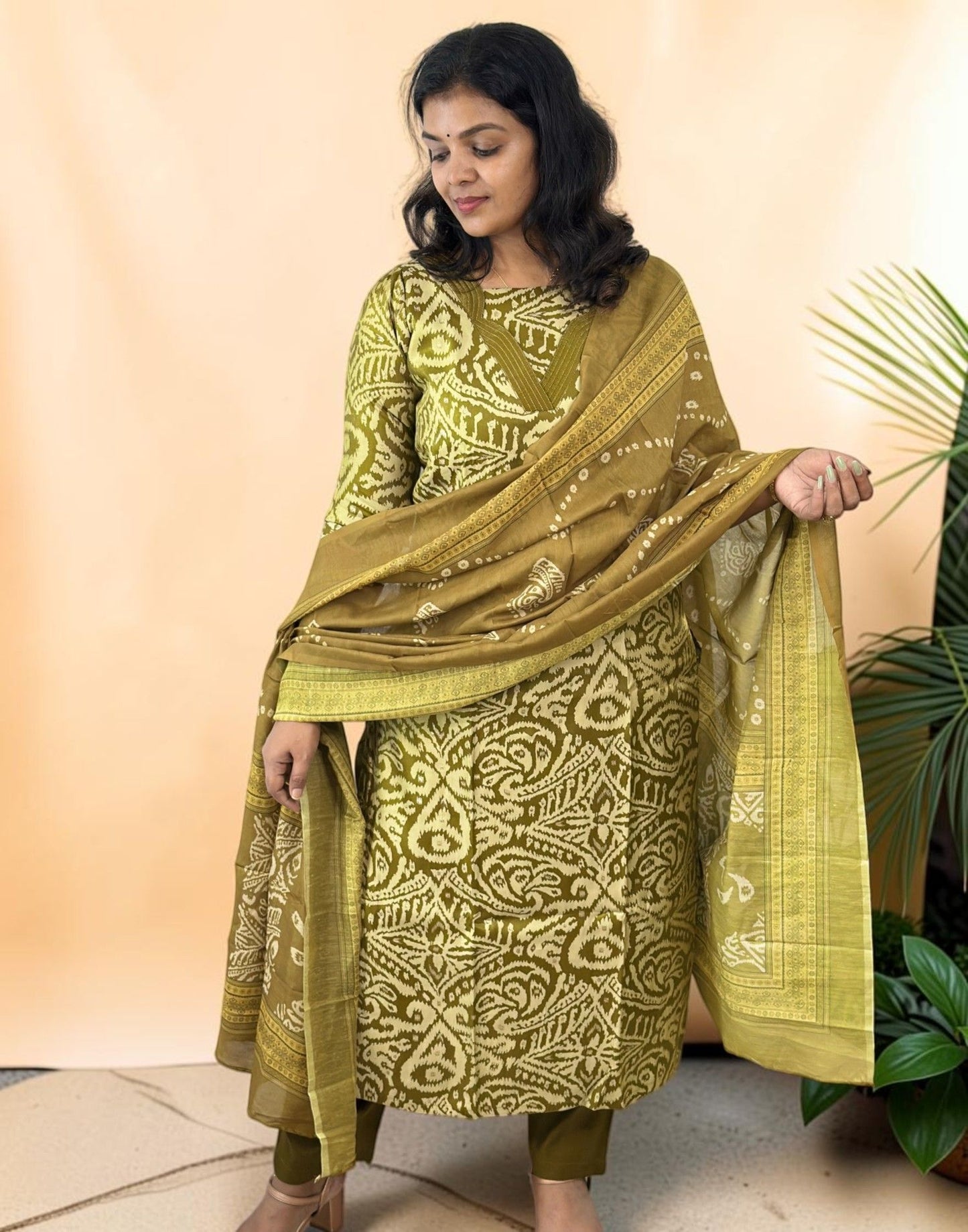 A woman models a three-piece olive green salwar kameez suit with white block prints and a matching dupatta, set against a warm beige backdrop with a decorative plant on the side.