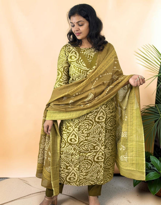 A woman models a three-piece olive green salwar kameez suit with white block prints and a matching dupatta, set against a warm beige backdrop with a decorative plant on the side.