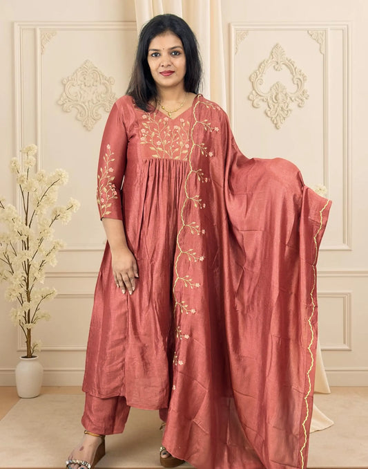 A woman models a dusty rose pink traditional Indian salwar kameez set with gold floral embroidery, featuring a matching dupatta draped over her shoulder, set against an elegant cream-colored paneled wall.