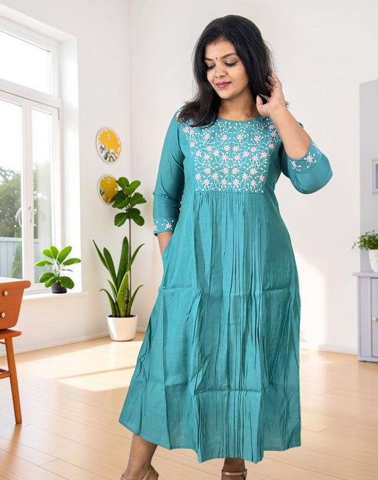 A woman modeling a teal green A-line kurta with white floral embroidery on the chest and sleeves, standing in a bright, modern room with light wood floors and potted plants.