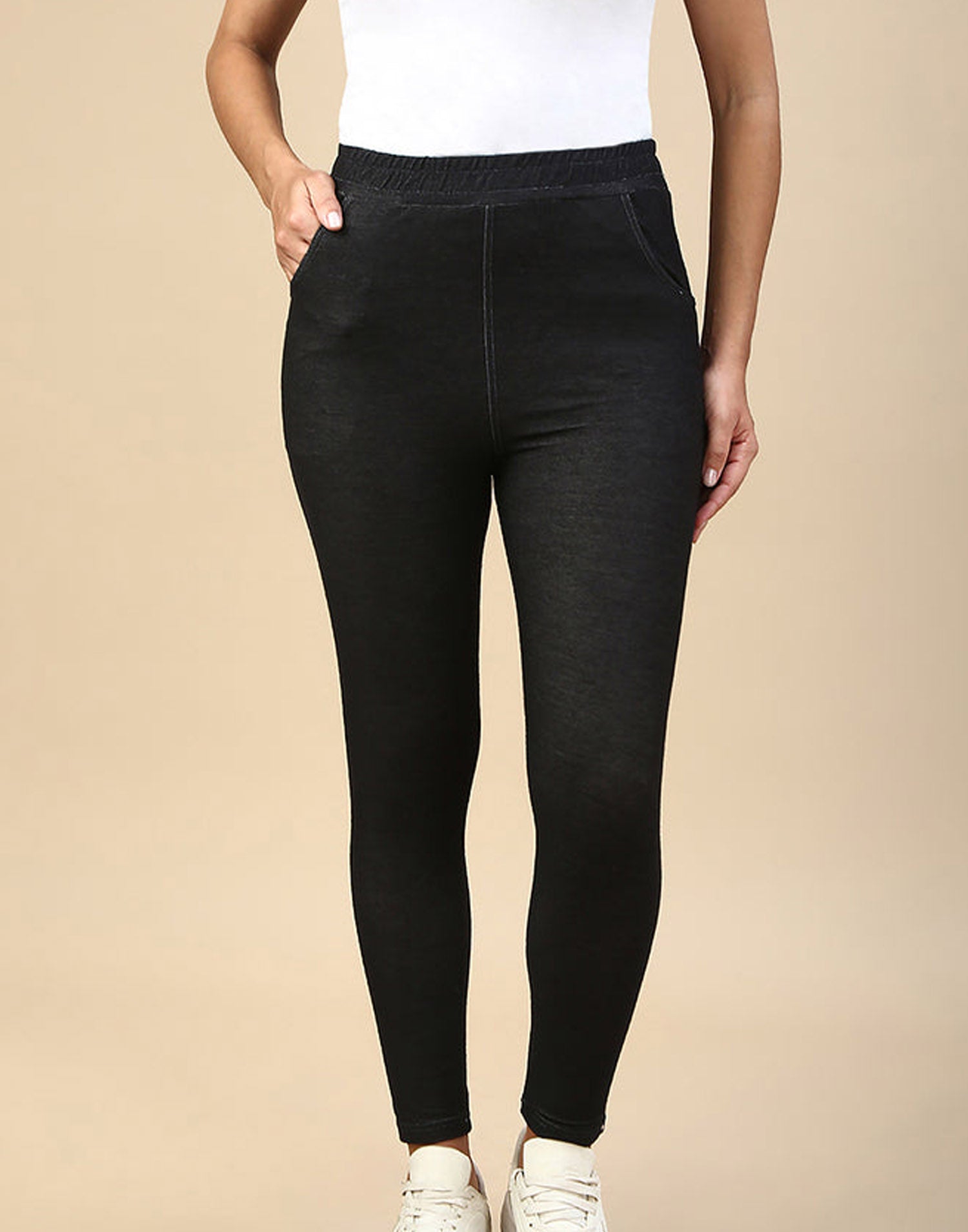 A model wearing black high-waisted leggings with side pockets, paired with a white top and white sneakers, set against a solid beige background.