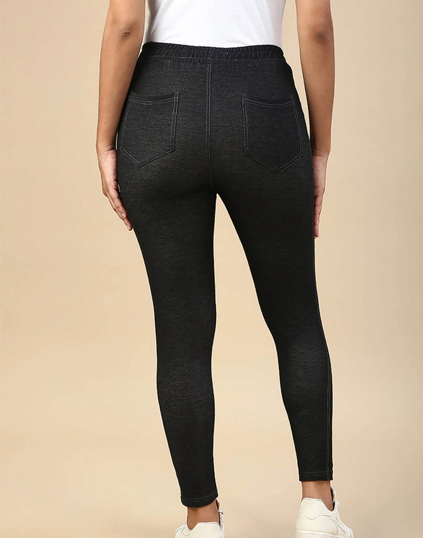 Rear view of a person wearing dark charcoal gray heathered leggings with a wide elastic waistband and two back pockets, paired with a white top and white sneakers, against a solid beige background.