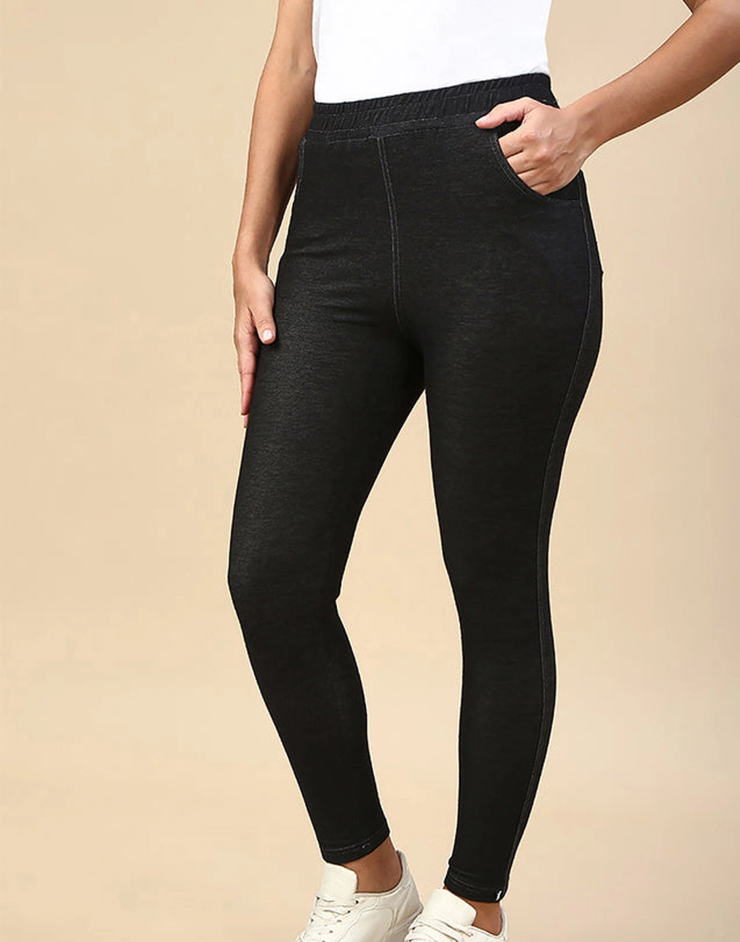 A person wearing black high-waisted leggings with side pockets, paired with a white top and white sneakers, shown against a plain beige studio background.