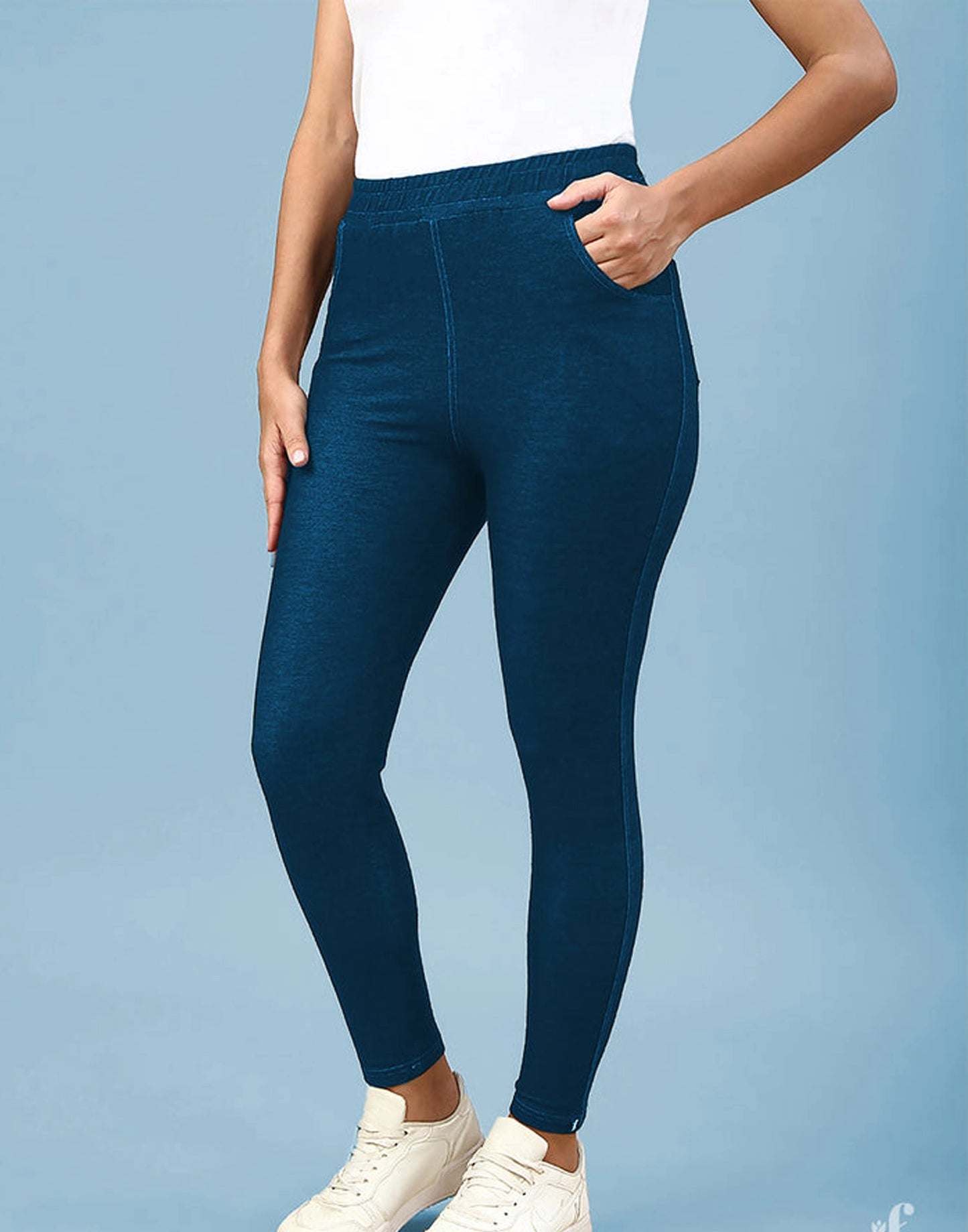 A model wearing dark teal blue high-waisted leggings with side pockets, paired with a white tank top and white sneakers, set against a solid light blue background.