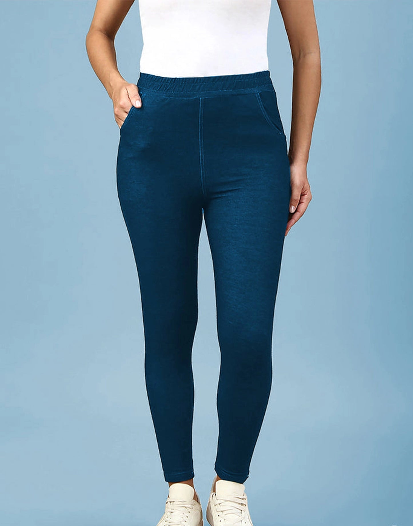 A person wearing dark teal blue leggings with a white top and white sneakers, standing against a solid light blue background.